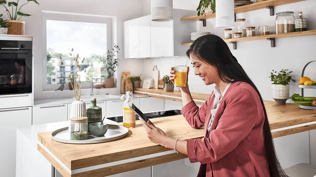 Femme buvant un jus à l'îlot d'une cuisine en U blanche avec coin repas bois Femme buvant un jus à l'îlot d'une cuisine en U blanche avec coin repas bois